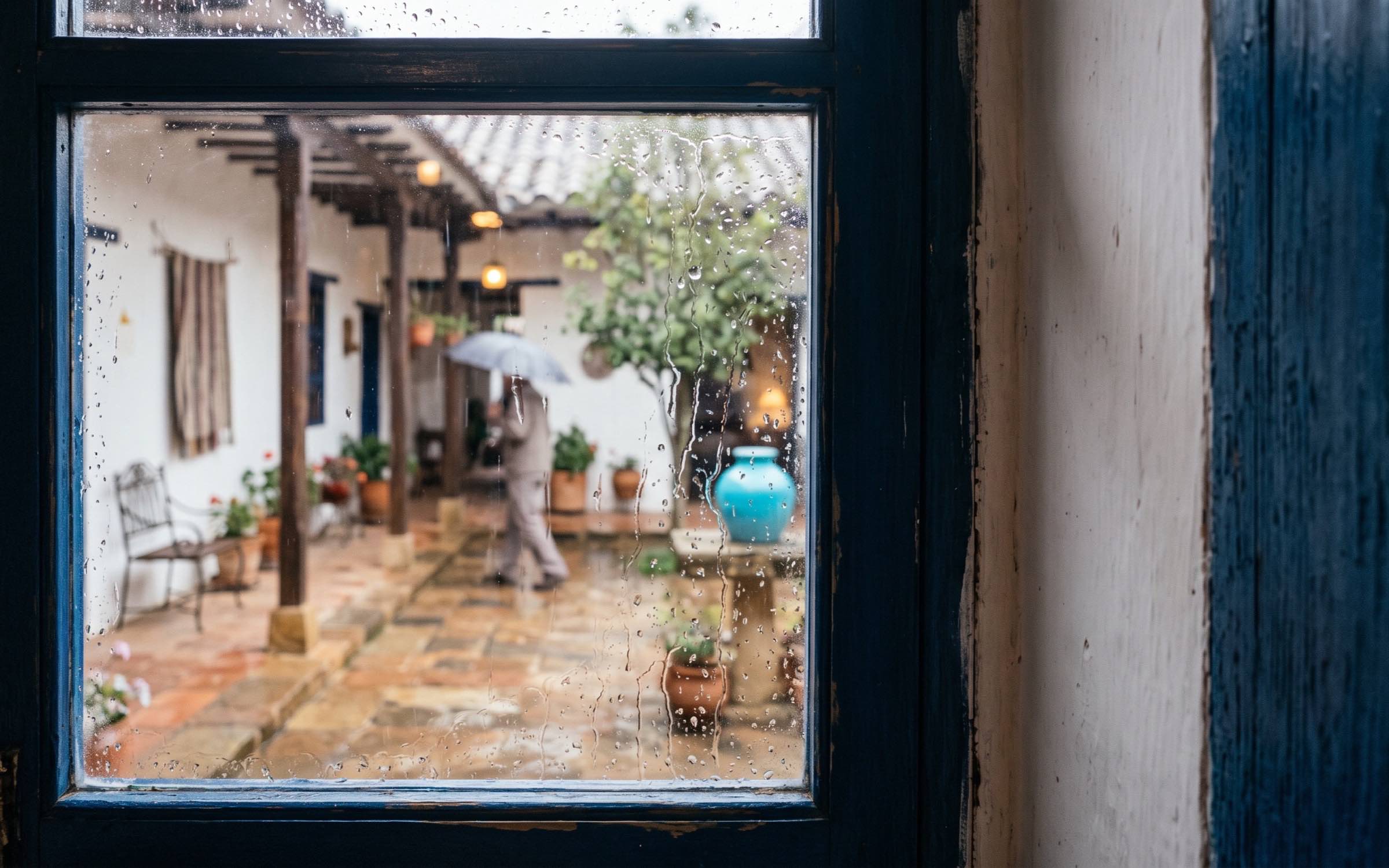 Vista de una ventana con gotas de lluvia y, al fondo, un patio de una casa colombiana ligeramente desenfocado.